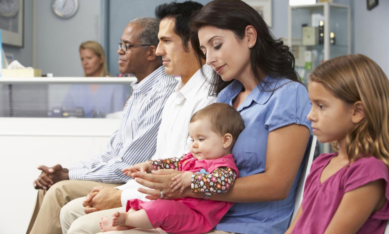 Diverse adults sitting in order inside a waiting room, a woman hold an infant. Next to her a child sits showing no emotion