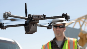 Close-up of a drone in flight near a construction worker wearing a safety vest and helmet.