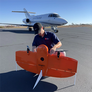 TDOT Technician inspecting an orange drone on an airport tarmac with an airplane parked in the background.