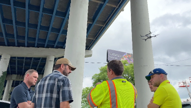 Group of workers standing beneath a highway overpass while a drone flies near the bridge structure above.