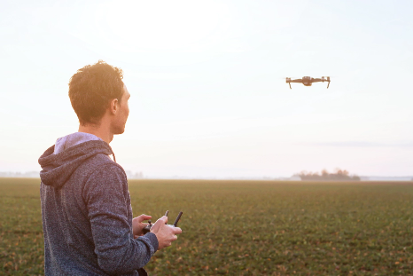 Person flying a drone over an open field during daylight using a handheld remote controller.