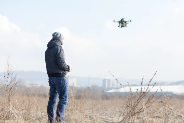 Person flying a drone in a dry, open field on a cold day, with buildings visible in the distant background.