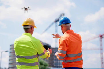 Two construction workers in safety gear flying a drone near a building site with cranes in the background.