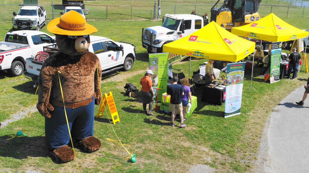 2023 Sevier County Wildfire Expo An outdoor fire safety event featuring a large inflatable Smokey Bear, yellow tents with fire prevention info, and fire trucks.