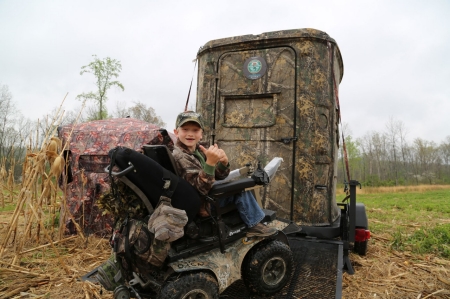 Young child gives thumbs up from his wheelchair when entering a hunting blind.