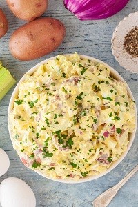 a large bowl of potato salad with whole potatoes and a red onion on the table next to it