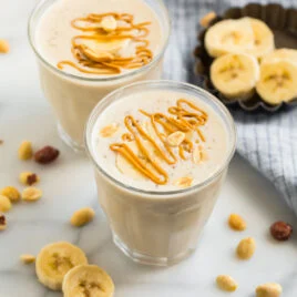 Peanut Butter Banana Smoothie two glasses of peanut butter banana smoothie with peanut butter drizzled on top; slices of banana are on the counter
