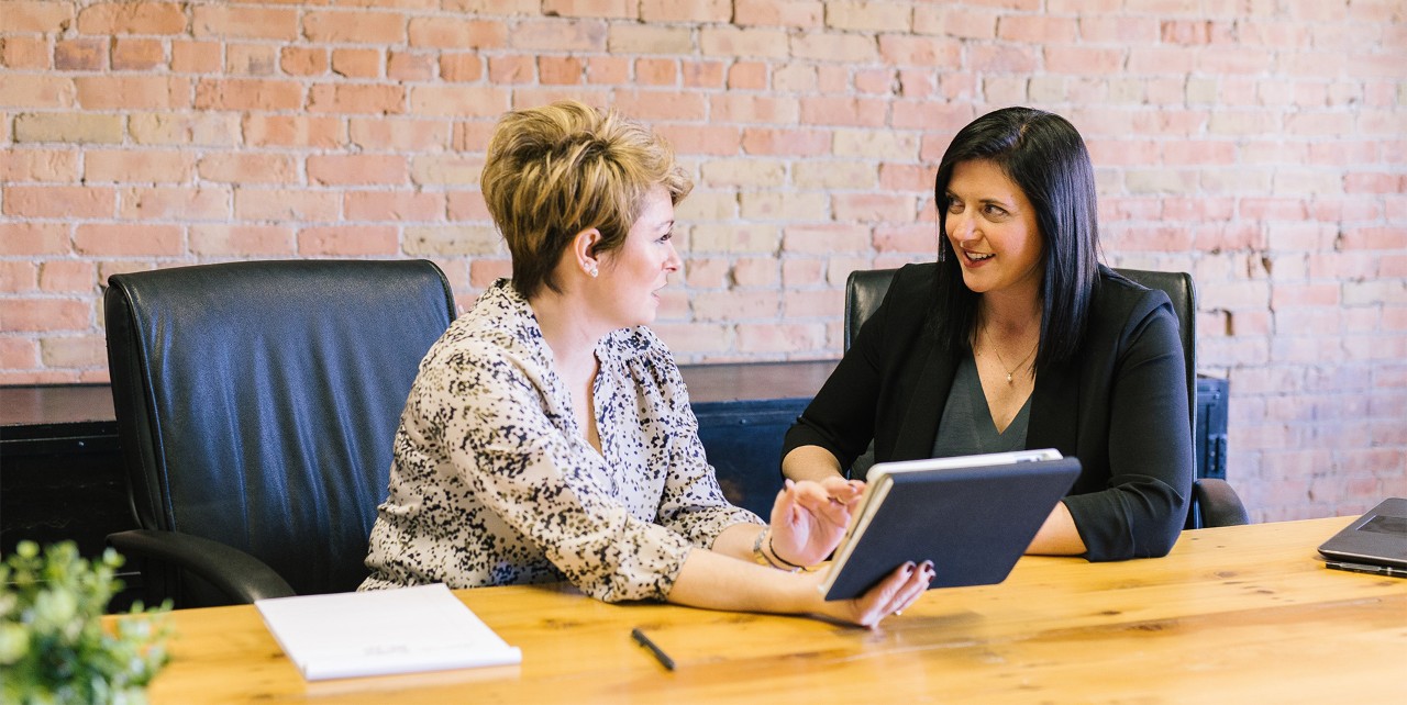 Two business women setting at a large conference room desk, working together.