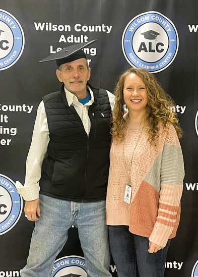 Man wearing a mortarboard stands next to a woman with a banner that says Wilson County Adult Learning Center in the background