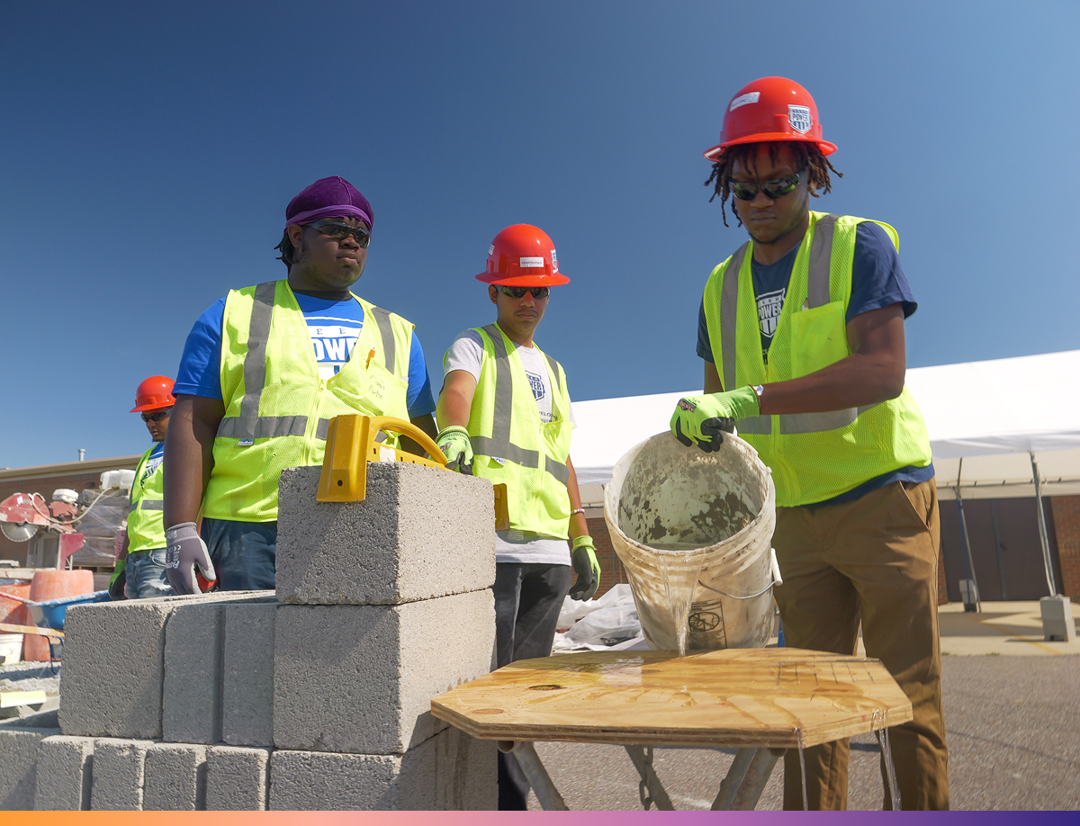 Youth Employment Program Young man wearing a helmet lifts a bucket next to a pile of bricks while two others watch